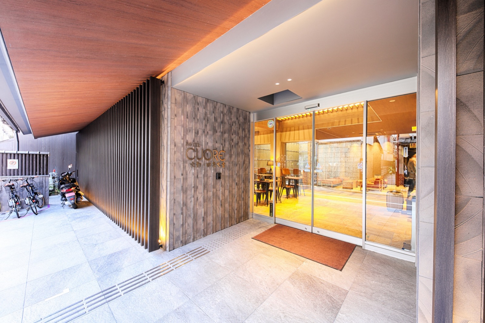 ホテルのエントランスアプローチ。木格子の壁とHOTEL CUOREのサイン、脇に駐輪スペース